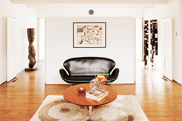 A lithograph by Philip Guston hangs above a Fritz Hansen settee and table; beyond them is the foyer, home to a tiki statue and a 1960s constructivist metal sculpture.