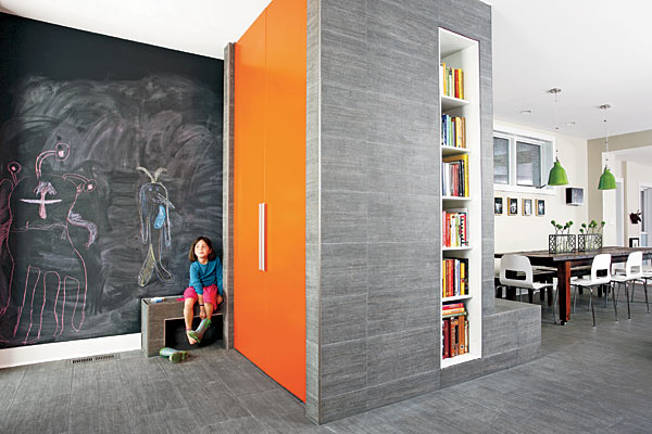Oversized gray porcelain tile covers a structure near the back door that houses a closet, a built-in bookcase, and a fireplace (not visible here) facing the kitchen table. Floors are clad with the same material. The bench in front of the chalkboard wall (with drawings by seven-year-old Alya) wraps around the structure and continues as a fireplace ledge.