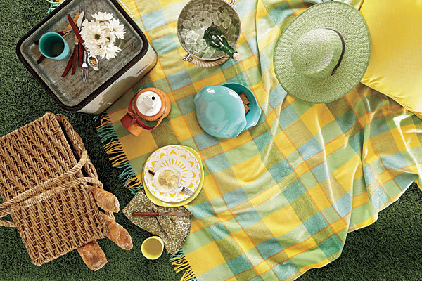 Russel Wright melamine tumbler, vintage wooden-handled flatware, vintage cooler, leather-wrapped thermos, polished nickel and brass ice bucket, vintage plaid wool blanket, outdoor acrylic pillow, Russel Wright melamine covered bowl, melamine plate, Russel Wright yellow melamine dinner plate, cotton napkin, and wicker picnic basket