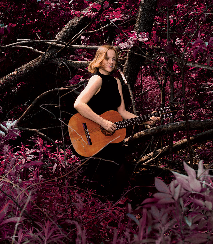 Jessica Risker with a guitar, standing in the middle of a group of trees with red leaves