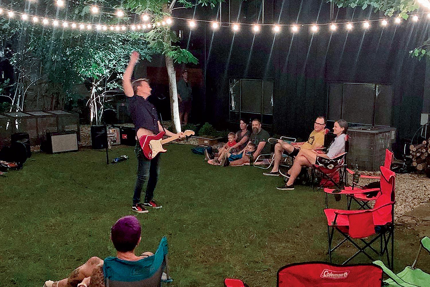 Jason Narducy playing guitar for a group of people in a backyard lit by outdoor string lights.